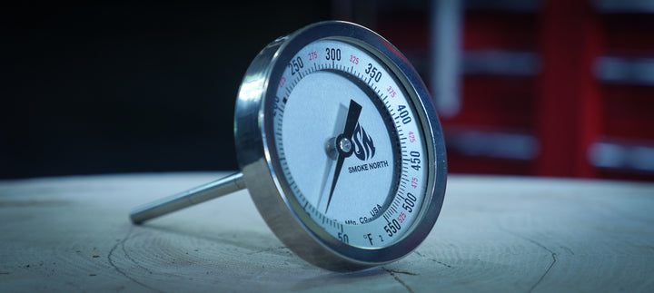 A stainless steel Tel Tru thermometer with the custom 'Smoke North' logo is shown sitting on a tree stump within a metal fabrication shop.